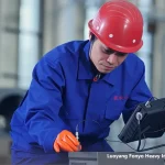 Worker in blue uniform and red helmet performing non-destructive testing on a metal component using a portable inspection device