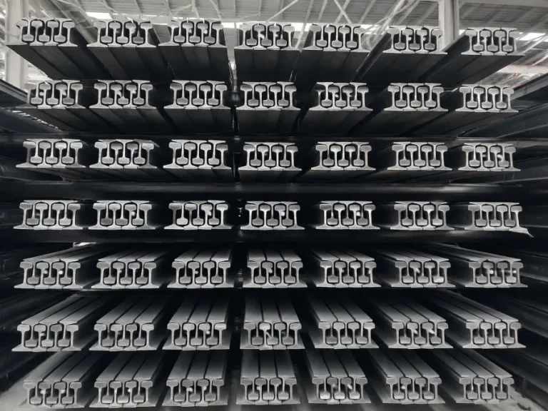 Neatly stacked steel rails sections stored in an industrial warehouse, showing rows of clean, unrusted railway tracks arranged in a symmetrical storage system.