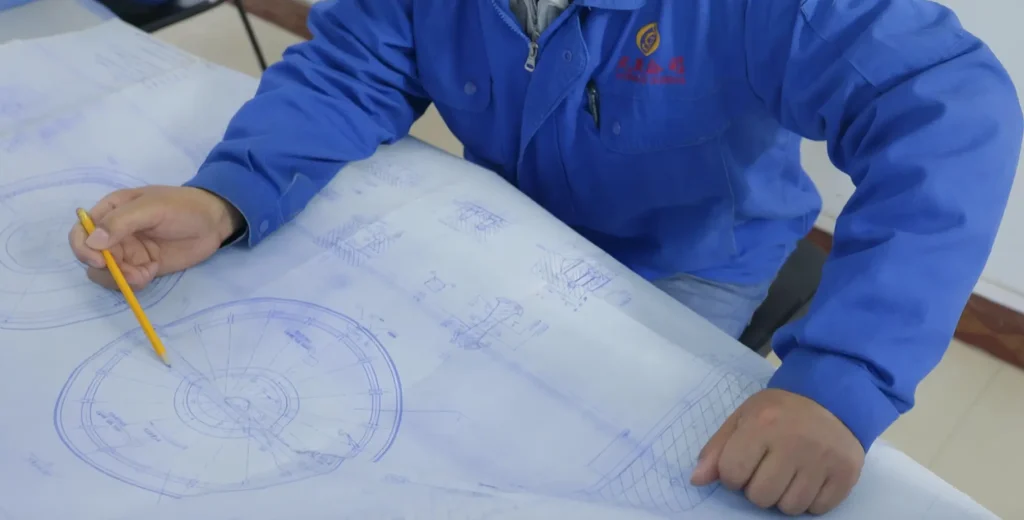High Manganese Steel Castings for Mining Wear Parts: A Buyer's Guide 3 A close-up view of an engineer of Fonyo in a blue work uniform examining large-scale technical blueprints spread on a table. The engineer holds a yellow pencil, pointing to a detailed circular mechanical drawing, likely verifying design specifications or dimensions.